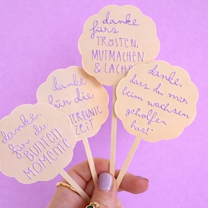 May include: Four light-coloured, cloud-shaped paper cutouts on wooden sticks, each with handwritten purple text. The cutouts are held against a purple background. The text includes phrases like "thank you" and "colourful moments."