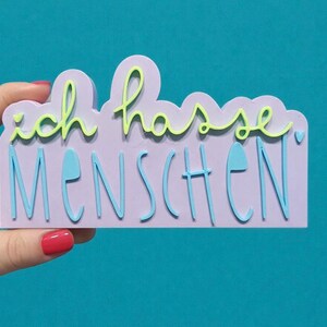 May include: A lavender-colored sign with the German phrase "ich hasse MENSCHEN" in light green and blue lettering. The sign is held by a hand with red nail polish against a teal background.