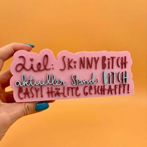 Può includere: Un oggetto rosa di forma rettangolare con le parole "Ziel: Skinny Bitch, aktueller Stand: Bitch, EASY! HÄLFTE GESCHAFFT!" in lettere bordeaux e azzurre. L'oggetto è tenuto da una mano con un'unghia turchese su sfondo arancione.