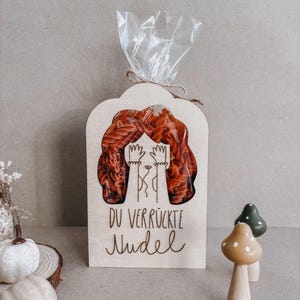 Könnte beinhalten: Eine Holzverpackung für Pasta mit einem transparenten Fenster, das rote Pasta zeigt. Der Holzrahmen hat ein ausgeschnittenes Design einer Person, die sich die Augen bedeckt. Der Text "DU VERRÜCKTE NUDEL" steht darunter.