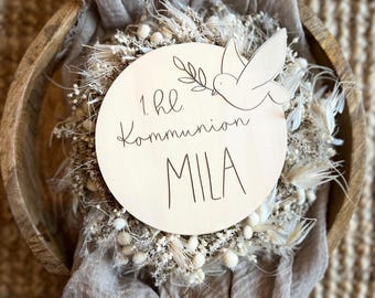 Pode incluir: Uma placa de madeira com o texto "1. hl. Kommunion MILA" e uma ilustração de pomba, rodeada por flores secas e um tecido bege. A placa está colocada numa tigela de madeira, criando uma estética rústica e natural.