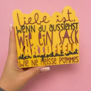 May include: A yellow sign with the German text "Liebe ist, wenn du aussiehst wie ein Sack Kartoffeln, aber angeschaut wirst wie ne Heisse Pommes" in brown and black lettering. The sign is held by a person.