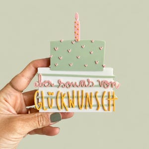 Könnte beinhalten: Ein dekoratives Schild in Kuchenform mit einer einzelnen Kerze. Der Kuchen ist salbeigrün mit kleinen rosa Herzen. Darunter befindet sich eine weiße rechteckige Basis mit dem rosa Schreibschrifttext "aber sowas von" und dem gelben Text "GLÜCKWUNSCH".