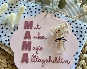 May include: A pink, cloud-shaped sign with the word "MAMA" in raised letters, along with other text and a small vase of dried flowers. The sign includes the words "Mut, Anker, Magie, Alltagsheldin" and a heart symbol.