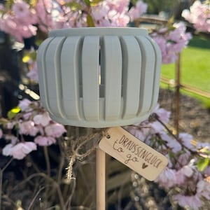 Può includere: Un portacandele cilindrico grigio chiaro con fessure verticali, attaccato a un bastoncino di legno. Una piccola etichetta di legno con il testo "DRAUSSENGLÜCK to go" è attaccata. Lo sfondo presenta fiori rosa.