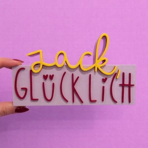 Könnte beinhalten: Ein rechteckiges Schild mit dem Wort "Jack" in gelber Schrift über dem Wort "GLÜCKLICH" in roter Schrift. Das Schild ist grau und hat einen lila Hintergrund.