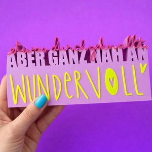 Puede incluir: Un letrero lavanda con las palabras "etwas durchgeknallt, aber ganz nah an Wundervoll" en rosa y amarillo. El letrero está sostenido por una mano con una uña pintada de azul sobre un fondo morado.