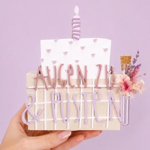 May include: A decorative rectangular block featuring a white cake topper with small hearts and a candle, displaying the German text AUGEN ZU & PUSTEN in raised lettering. A small glass vial with a cork stopper and dried flowers is attached to the side.
