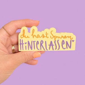 May include: A yellow sticker with the German phrase "du hast Spuren hinterlassen" in orange and purple lettering. The sticker is held against a purple background.