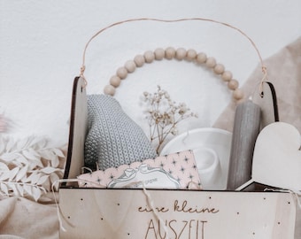 May include: A wooden gift box with a handle, containing a gray knitted item, a white teacup, a candle, and a wooden heart. The box has the text "eine kleine AUSZEIT für dich" and is decorated with small dots.