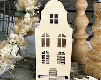 May include: A light-colored wooden house-shaped candle holder with cut-out windows. The holder has a rectangular base with a drawer and is surrounded by dried flowers and wooden candle holders.