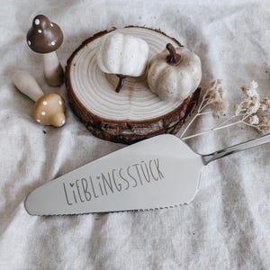Puede incluir: Una composición decorativa con una espátula plateada para tartas con el texto "LIEBLINGSSTÜCK", dos figuras de setas y dos pequeñas calabazas sobre una rodaja de madera. Las calabazas son blancas y beiges, y las setas son marrones y beiges.
