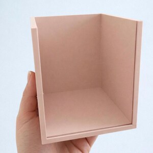 Könnte beinhalten: Ein kleiner, quadratischer Behälter mit offener Oberseite in einem gedämpften Rosaton. Der Behälter wird in einer Hand gehalten und zeigt seine klaren Linien und sein schlichtes Design. Die Innen- und Außenflächen sind gleichfarbig.