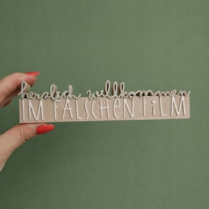 May include: A wooden sign with the words "herzlich willkommen IM FALSCHEN FILM" in white script. The sign is held against a green background.