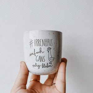 Może przedstawiać: Nakrapiany ceramiczny kubek trzymany w dłoni. Kubek ma czarny tekst i ilustrację gęsi. Tekst brzmi "#IRRENHAUS einfach GANS ruhig bleiben". Kubek jest jasnoszary z małymi ciemnymi plamkami.