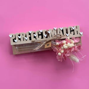 Könnte beinhalten: Ein rechteckiges Holzschild mit dem Schriftzug PERFECT MATCH in erhabenen Buchstaben, verziert mit kleinen weißen Herzen. Ein durchsichtiges Glas-Reagenzglas ist vorne befestigt und mit einer Juteschleife, einem kleinen Herzanhänger und rosa Trockenblumen dekoriert.
