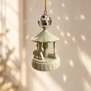 Può includere: Un ornamento a giostra sospeso con finitura verde salvia opaco, caratterizzato da un cavallo scolpito su una base circolare con tetto smerlato. Una piccola palla da discoteca argentata riflettente è fissata in alto, appesa a un sottile filo dorato.