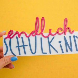Könnte beinhalten: Ein weißes rechteckiges Schild mit den deutschen Wörtern "endlich SCHULKIND" in Blau und Pink. Das Wort "endlich" ist in rosa Schreibschrift, und "SCHULKIND" ist in blauen Großbuchstaben. Das Schild wird vor einem gelben Hintergrund gehalten.