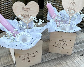 May include: Two wooden gift boxes with heart-shaped toppers that read "Happy Birthday." Each box contains a pink candle, a white decorative item, and white flowers. One box says "Heute ist dein Tag" and the other says "oh HAPPY day."