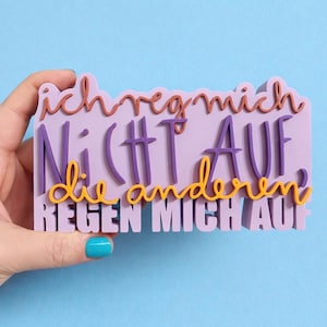Puede incluir: Un letrero rectangular de color lavanda con la frase alemana "Ich reg mich nicht auf die anderen regen mich auf" en varios colores. El texto está en una mezcla de fuentes y estilos, sostenido por una mano.
