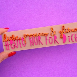 Könnte beinhalten: Ein rechteckiges, hellbraunes Schild mit den Worten "liebe prosecco & glitzer" und "#HEUTE NUR FÜR DICH" in orangefarbenen und pinkfarbenen Buchstaben. Das Schild wird vor einem lila Hintergrund gehalten.