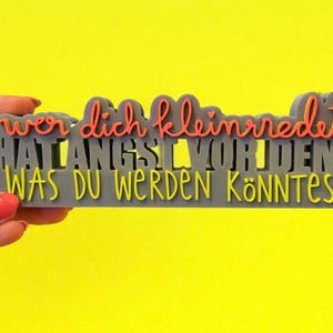Pode incluir: Uma placa retangular cinza com a frase alemã "wer dich kleinredet hat angst vor dem was du werden könntest" em letras laranja e amarelas. A placa é segurada contra um fundo amarelo brilhante.