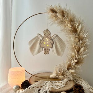 Op de afbeelding: Een houten engel ornament met een gouden halo en witte garen vleugels hangt aan een gouden metalen ring. De ring is versierd met gedroogd pampasgras en andere gedroogde bloemen. De engel wordt verlicht met een warm wit licht.