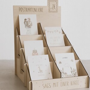 Könnte beinhalten: Ein Postkartenständer aus Holz mit mehreren Fächern, der verschiedene illustrierte Postkarten enthält. Der Ständer trägt die Aufschrift "POSTKARTENLIEBE" und "SAGS MIT EINER KARTE". Eine Briefmarkengrafik mit "2€" ist ebenfalls zu sehen.