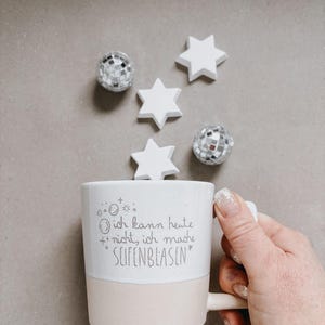 Może przedstawiać: Biały ceramiczny kubek z jasnoróżową podstawą, trzymany w dłoni. Na kubku widnieje napis "Ich kann heute nicht, ich mache SEIFENBLASEN". Rozrzucone są białe dekoracje w kształcie gwiazd i dwie ozdoby w kształcie kuli dyskotekowej.