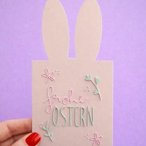 Puede incluir: Una tarjeta de Pascua rosa claro con orejas de conejo en la parte superior. La tarjeta presenta las palabras "Frohe Ostern" en escritura rosa y verde, junto con adornos florales. El fondo es morado liso.