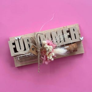 Op de afbeelding: Een rechthoekig houten bordje met de beige tekst FÜR IMMER aan de voorzijde bevestigd. Een horizontaal gemonteerde glazen reageerbuis met kurk bevat gedroogde roze en witte bloemen, vastgebonden met touw en een klein hartvormig label.