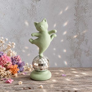 Könnte beinhalten: Eine matte, salbeigrüne Katzenfigur in tanzender Pose, die auf einer kleinen, silbernen Discokugel mit Spiegelfliesen balanciert und auf einem passenden, runden, salbeigrünen Sockel steht.