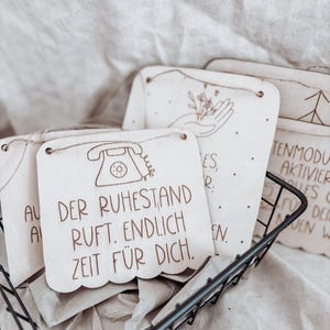 May include: A collection of wooden plaques with engraved designs and text, displayed in a black wire basket. The plaques feature illustrations of a telephone, hand holding flowers, and text in German. The text reads "DER RUHESTAND RUFT. ENDLICH ZEIT FÜR DICH."