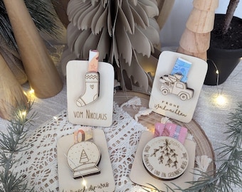 May include: Four wooden Christmas ornaments with different designs. The ornaments are decorated with a sock, a snow globe, a mistletoe sprig, and a truck carrying presents. The ornaments are on a wooden surface with a white lace tablecloth and a string of lights.
