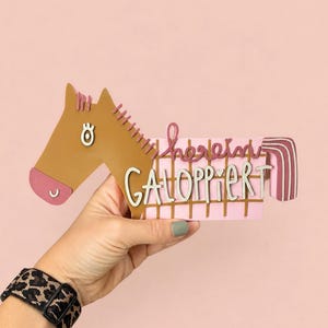 May include: A decorative wall sign shaped like a brown horse head with a pink muzzle and eye detail. The body features a pink grid pattern with the text "herein GALOPPIERT" in white and dark pink lettering, finished with a striped pink tail.