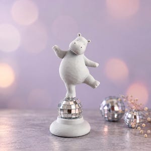 Könnte beinhalten: Eine hellgraue Nilpferd-Figur, die auf einem Bein auf einer kugelförmigen Discokugel mit Spiegelkacheln steht. Das Nilpferd hat eine glatte, matte Oberfläche und steht auf einem runden Sockel. Zwei weitere kleine Discokugeln sind daneben zu sehen.