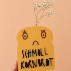 Könnte beinhalten: Eine gelbe, brotförmige Vase mit einem Cartoon-Gesicht und den schwarzen Worten "SCHMOLL KORNBROT". Die Vase enthält einen Zweig weißer Blumen. Der Hintergrund ist einfarbig pfirsichfarben.