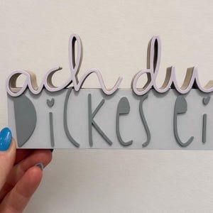 Könnte beinhalten: Ein graues rechteckiges Schild mit dem deutschen Satz "ach du dickes Ei" in grauen und lavendelfarbenen Buchstaben. Das Schild wird von einer Hand mit blau lackiertem Fingernagel gehalten.