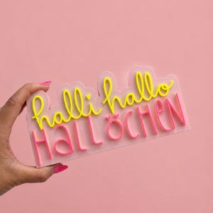 May include: A rectangular light pink sign featuring raised lettering. The top line displays the yellow cursive text halli hallo with a small heart. The bottom line features the bright pink block-letter text HALLÖCHEN with two small hearts above the O.
