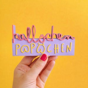 Könnte beinhalten: Eine Hand hält ein lila Schild mit den Worten "hallöchen P@PÖCHEN" in rosa und gelben Buchstaben. Das Schild befindet sich vor einem leuchtend gelben Hintergrund. Die Buchstaben sind in einer verspielten, skurrilen Schriftart.