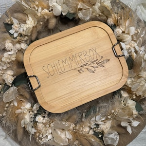 Könnte beinhalten: Eine natürliche Holz-Lunchbox mit einem schwarzen Verschluss und den Worten "Schlemmerbox" auf dem Deckel graviert.