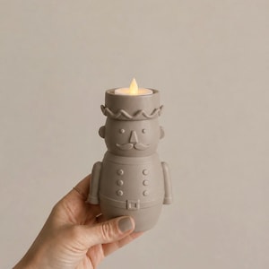 May include: A matte taupe ceramic nutcracker candle holder shaped like a soldier with a cylindrical hat, a mustache, and a uniform with buttons and a belt. The top of the hat features a small, glowing, warm white LED tea light candle.