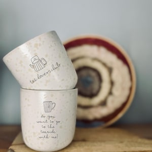 Op de afbeelding: Twee crèmekleurige keramische mokken met een gespikkeld ontwerp. Eén mok heeft een theepotillustratie en de tekst "tee lovers club". De andere mok heeft een theekopillustratie en de tekst "do you want to go to the seaside with me!". Een decoratieve schaal staat op de achtergrond.