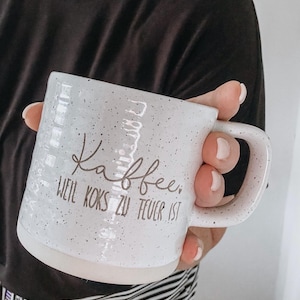 Könnte beinhalten: Weiß gesprenkelter Keramikbecher mit dem Wort "Kaffee" und dem Satz "Weil Koks zu teuer ist" in brauner Schrift. Der Becher hat einen großen Henkel und einen weißen Sockel.