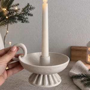 Könnte beinhalten: Ein matter, cremeweißer Kerzenhalter aus Keramik mit einer runden Schale, einem geriffelten Sockel und einem kleinen seitlichen Griff. Der Halter enthält eine einzelne, lange, cremefarbene Stabkerze mit einer leuchtenden, flammenförmigen LED-Spitze.