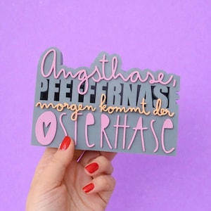 May include: A grey sign with pink and orange lettering. The sign reads "Angsthase, Pfeffernase, morgen kommt der OSTERHASE" in a playful font. A heart shape is included in the design. The sign is held against a purple background.