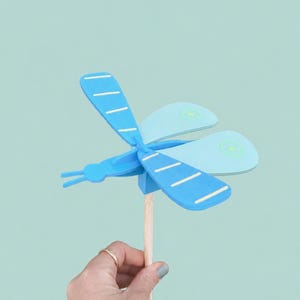 Op de afbeelding: Een blauwe en lichtblauwe houten libelle speelgoed met witte strepen op de vleugels. De libelle is bevestigd aan een houten stok. De achtergrond is lichtblauwgroen.