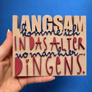 May include: A wooden sign with the German phrase "LANGSAM komme ich IN DAS ALTER wo man hier... DINGENS." The letters are in a mix of colors, with the main text in a natural wood tone, and the other text in red and blue.