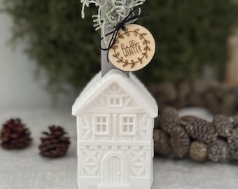 May include: A white candle in a house-shaped holder, with a gray candle and a wooden tag that says "Hello Winter". The candle is decorated with greenery and a black ribbon. Two pinecones are in the background.