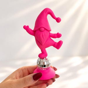 Könnte beinhalten: Eine leuchtend neonpinke Zwergenfigur mit spitzem Hut und langem Bart, die in einer tanzenden Pose dargestellt ist. Der Zwerg ist auf einer kleinen, kugelförmigen silbernen Discokugel mit reflektierender Oberfläche und einem pinken Sockel montiert.